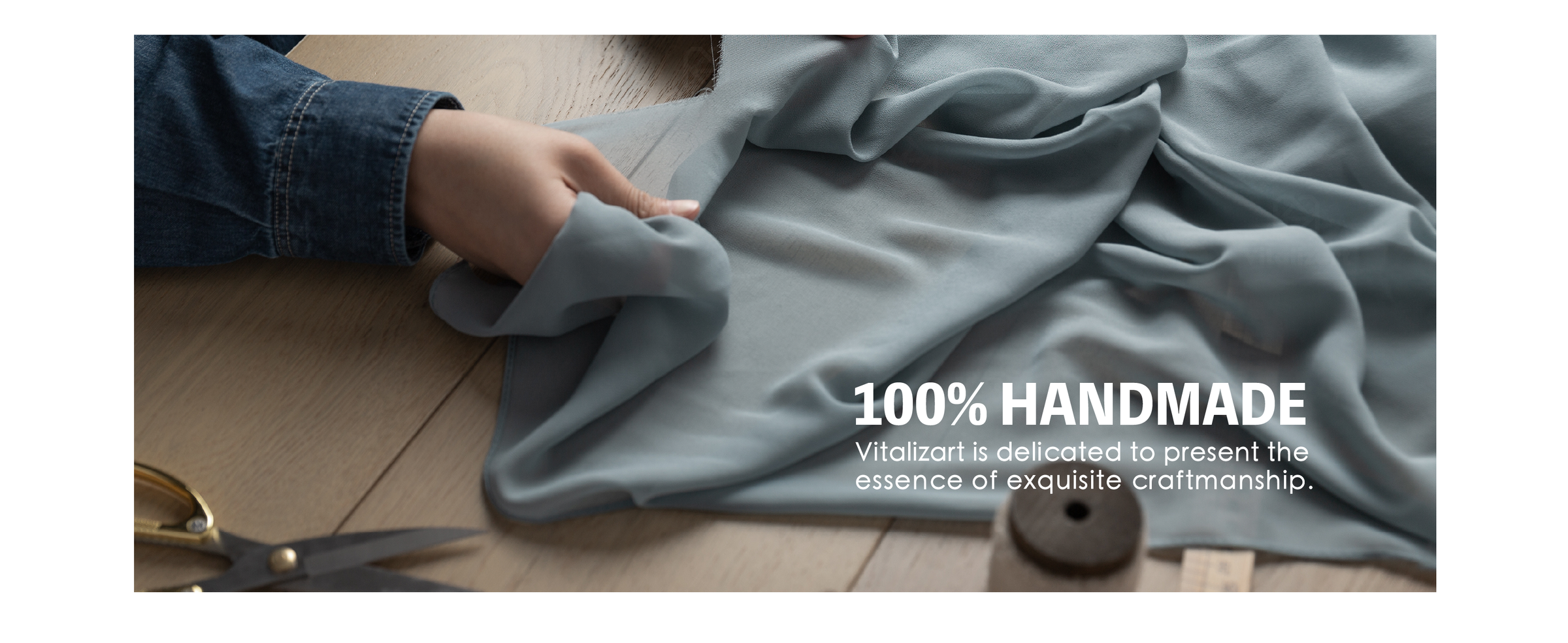 Hands gathering blue-gray fabric on a worktable beside tailor's shears, a spool, and a ruler.