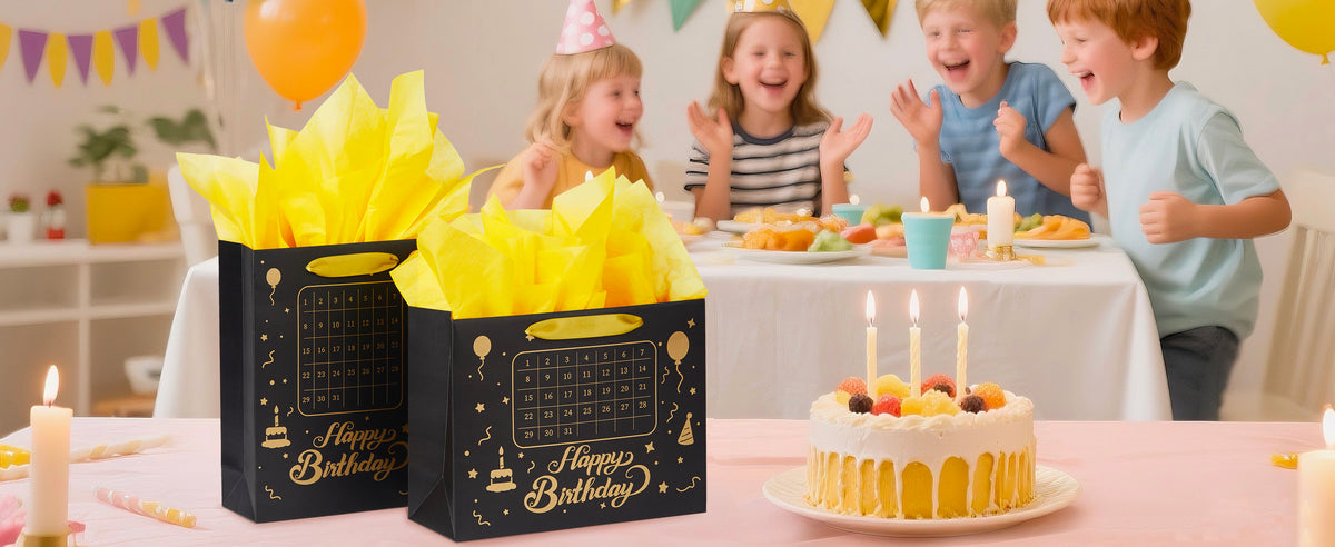 Two black calendar birthday gift bags with gold handles and yellow tissue beside a candlelit cake