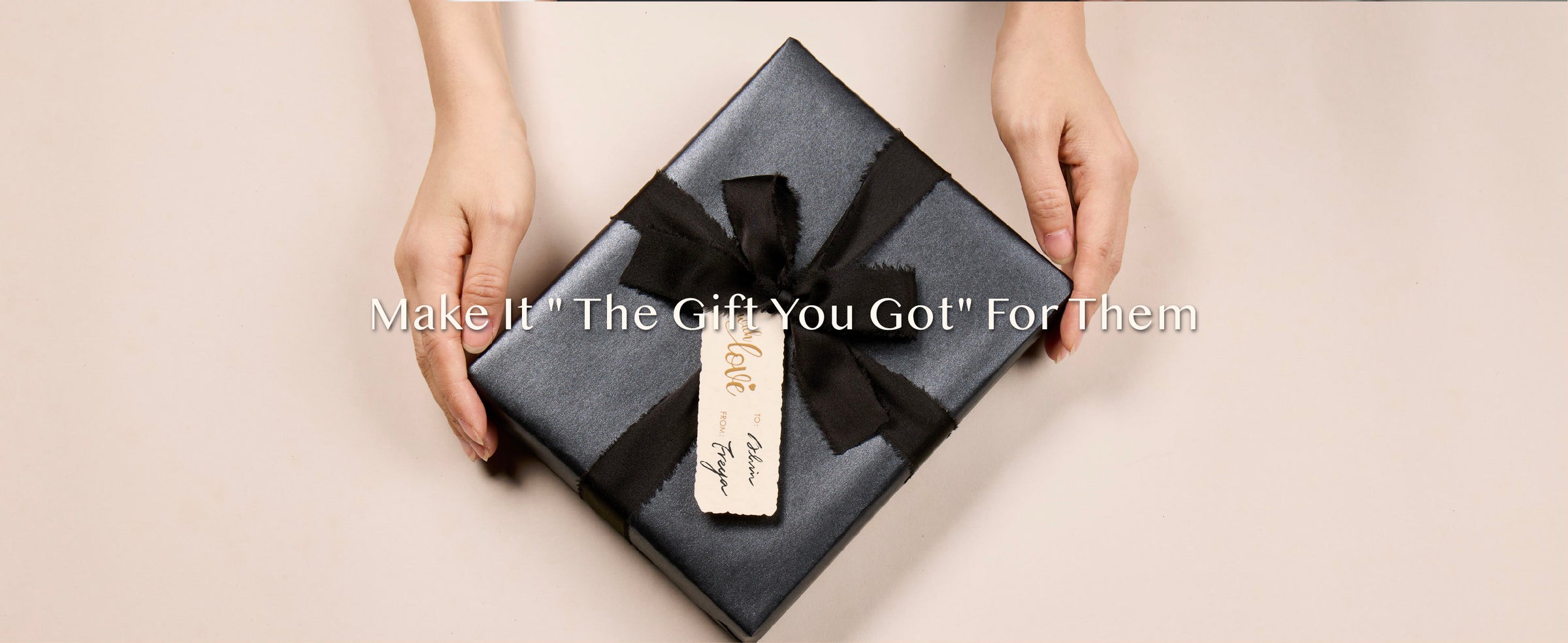 Hands presenting a square gift wrapped in pearly charcoal paper with a black bow and cream tag