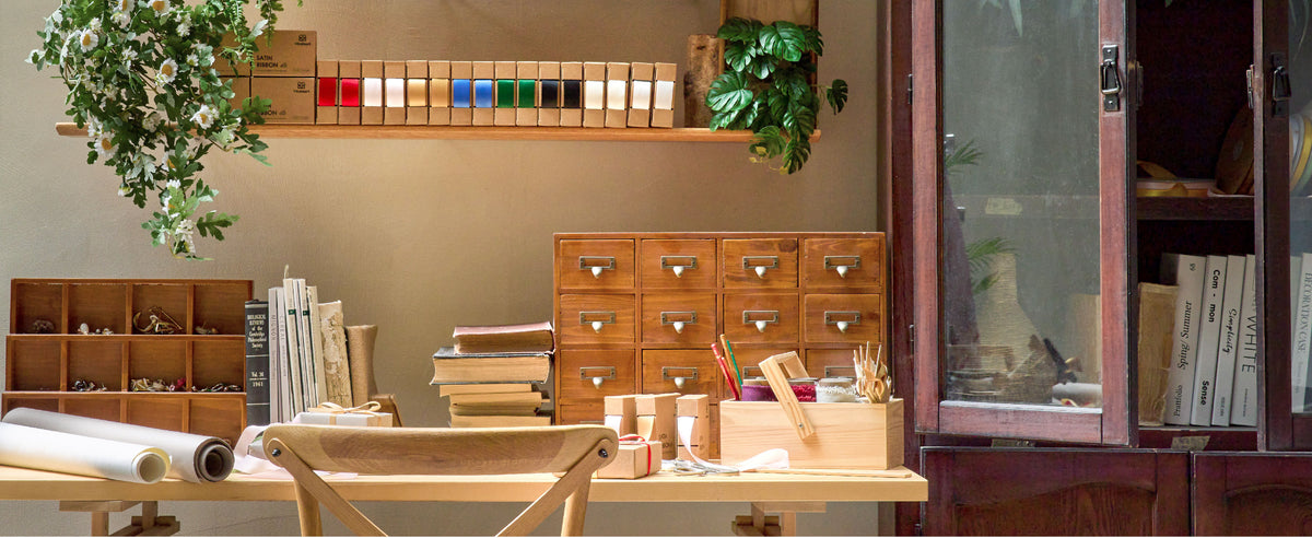 Sunlit craft studio with assorted ribbon dispensers, books, scissors, and supplies on a wooden worktable.