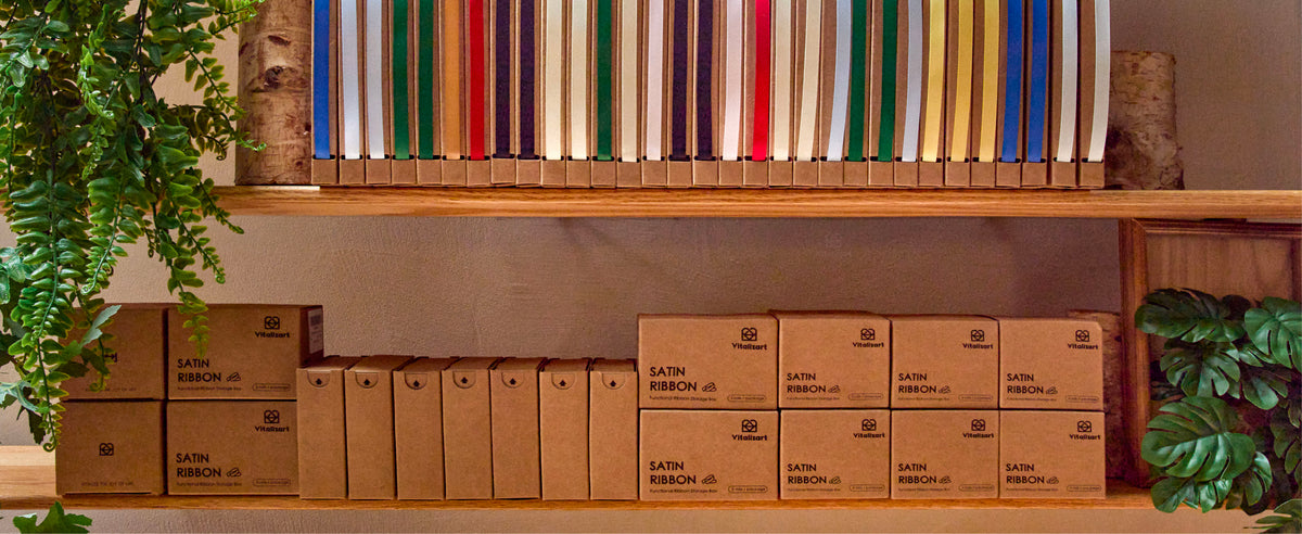 Assorted colorful satin ribbons stand in slim kraft dispensers on wooden craft-room shelves.