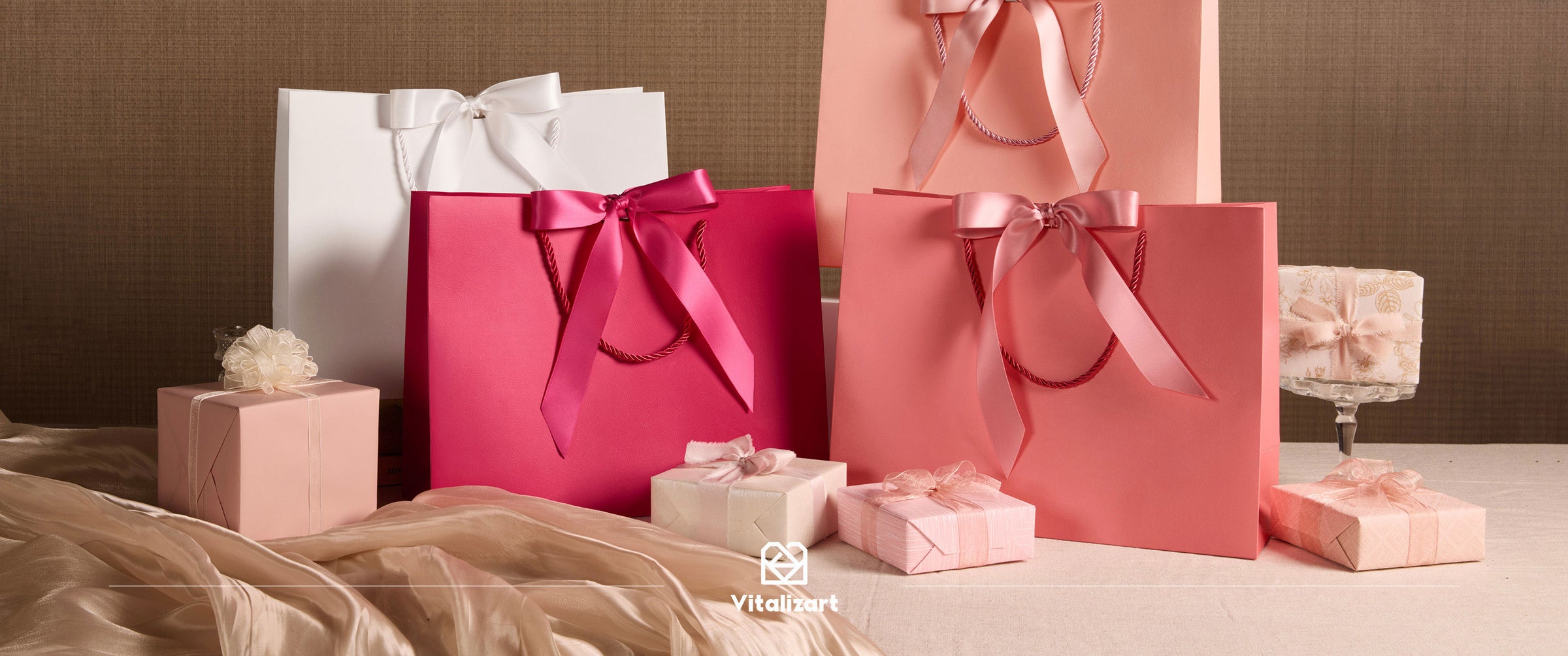 Four white and pink paper gift bags with satin bows displayed with wrapped gifts and draped fabric