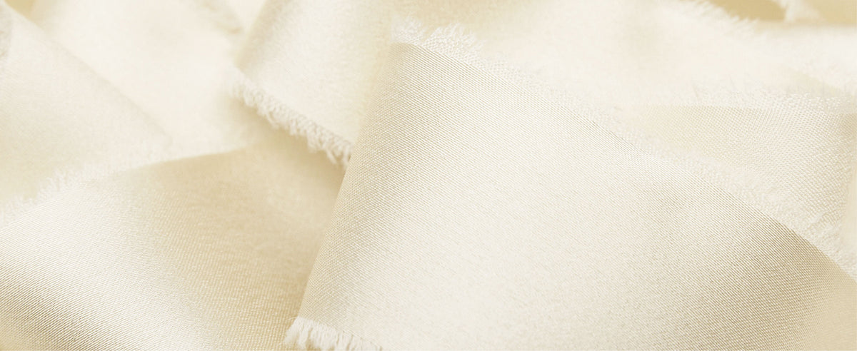 Close-up of ivory satin ribbon in soft overlapping folds, showing its sheen, fine weave, and frayed edges.