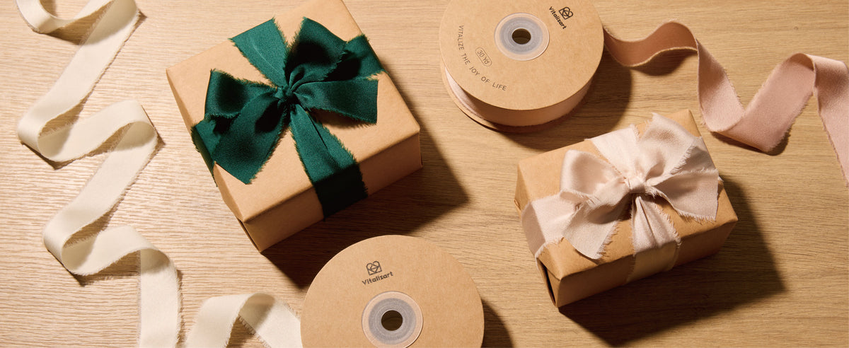 Green and blush frayed satin ribbon bows on kraft gifts beside ribbon spools on a wooden table.