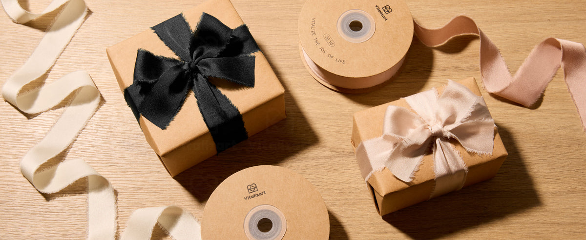 Black and blush frayed satin ribbon bows on kraft gift boxes beside ribbon spools on a wood table.