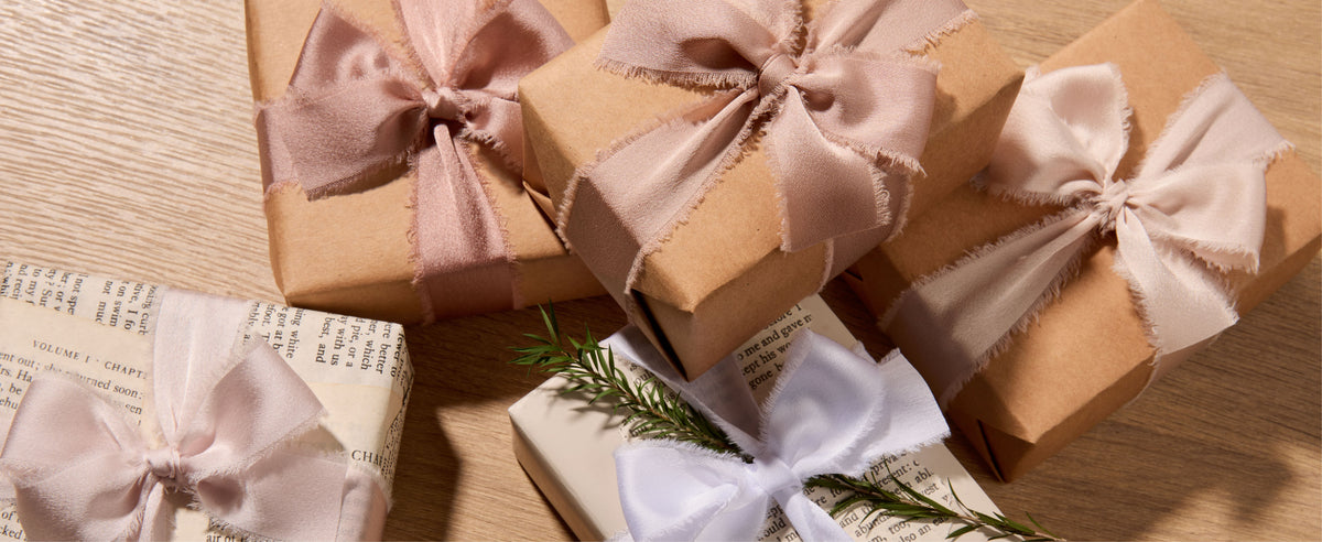 Kraft and white gift boxes tied with nude, ivory, and white frayed satin ribbon bows.
