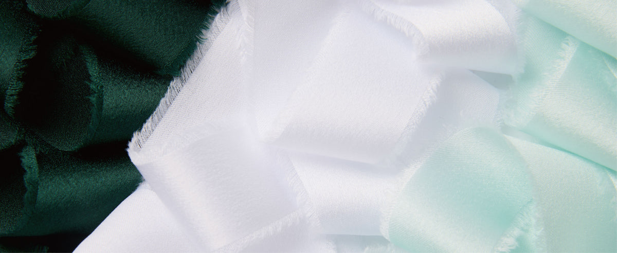 Close-up of folded frayed satin ribbons in dark green, white, and pale mint with a soft sheen.