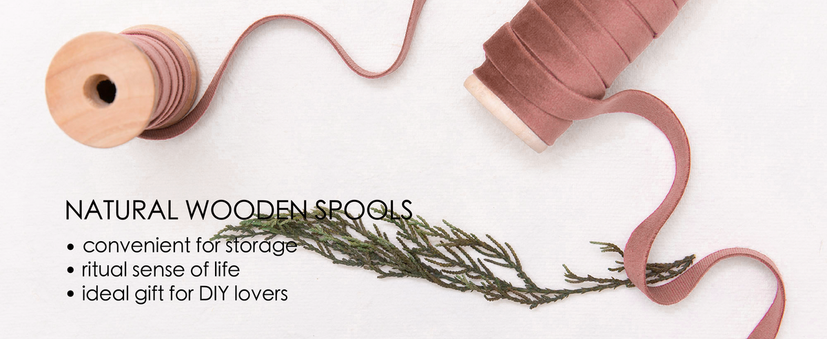 Rose-colored velvet ribbon unwinds between two natural wooden spools on white fabric with greenery.
