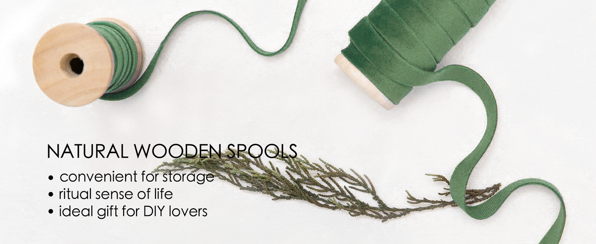 Green velvet ribbon unwinds between two natural wooden spools on white fabric with greenery.