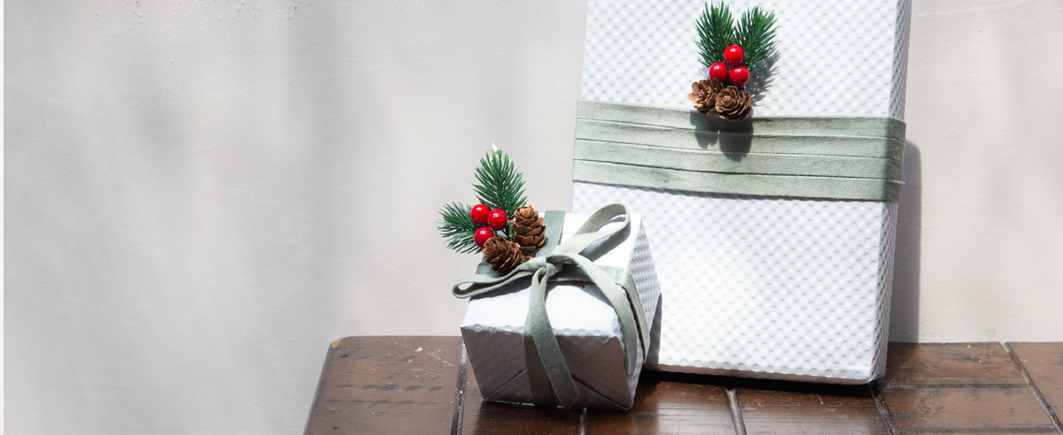 White holiday gifts wrapped with dusty green velvet ribbon, pinecones, evergreen sprigs, and red berries.