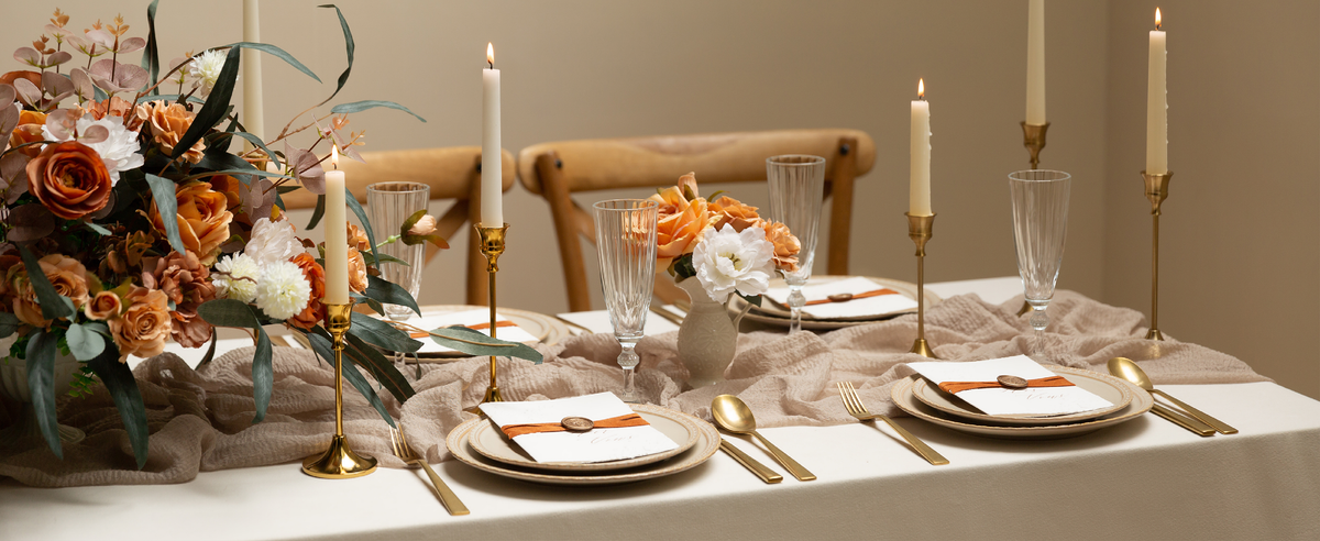 Terracotta ribbon accents formal place settings with gold flatware, flowers, and lit taper candles.