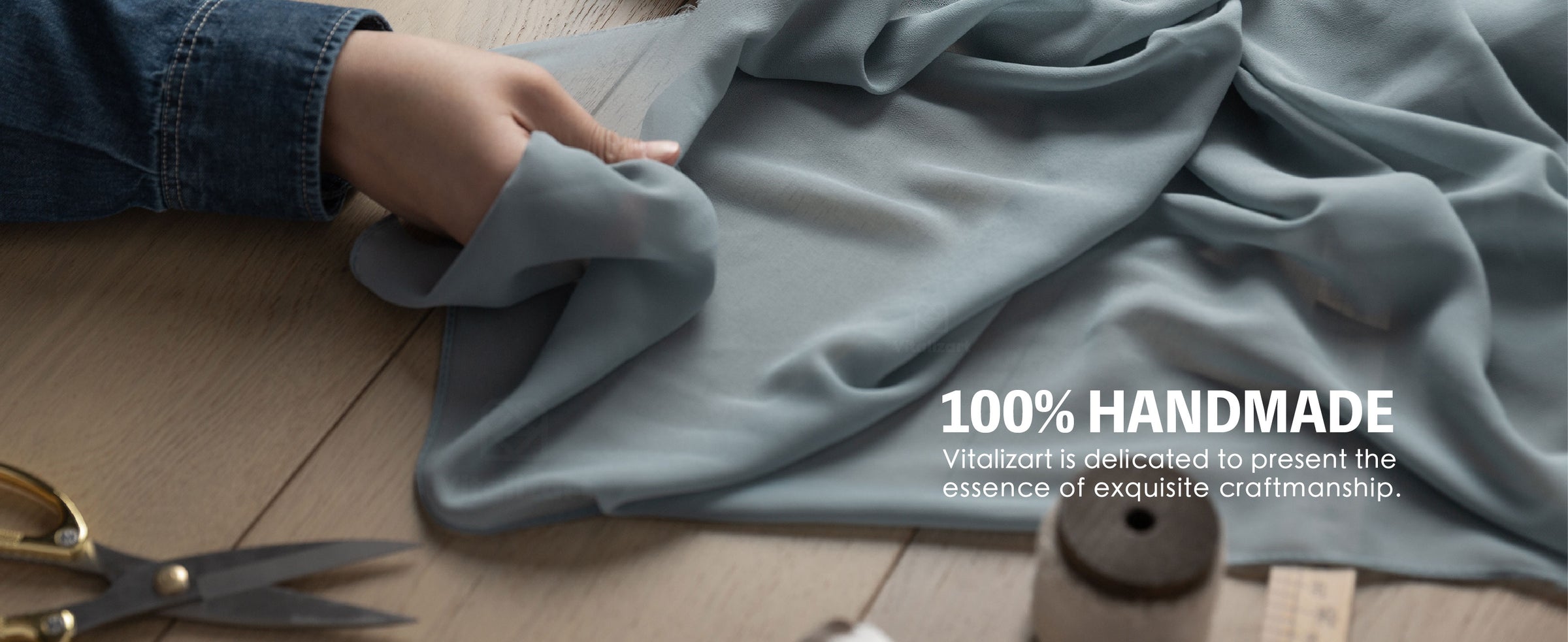Craftsperson handling flowing gray fabric on a worktable with scissors, ruler, and spool