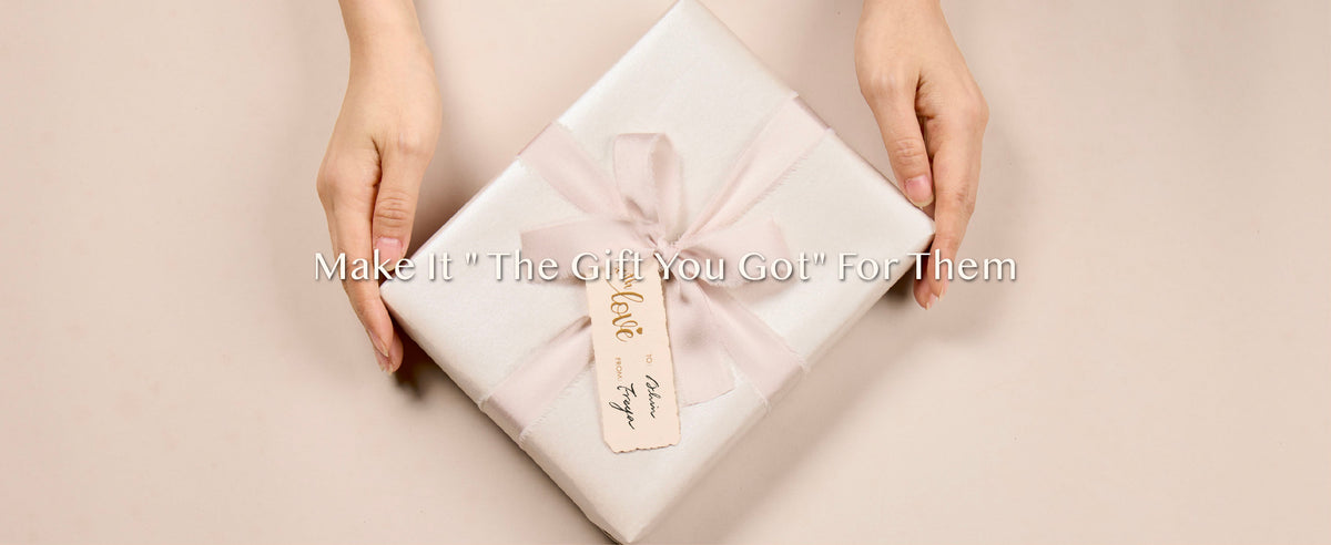 Hands holding a square gift in pearly white wrapping paper with a light nude bow and cream tag