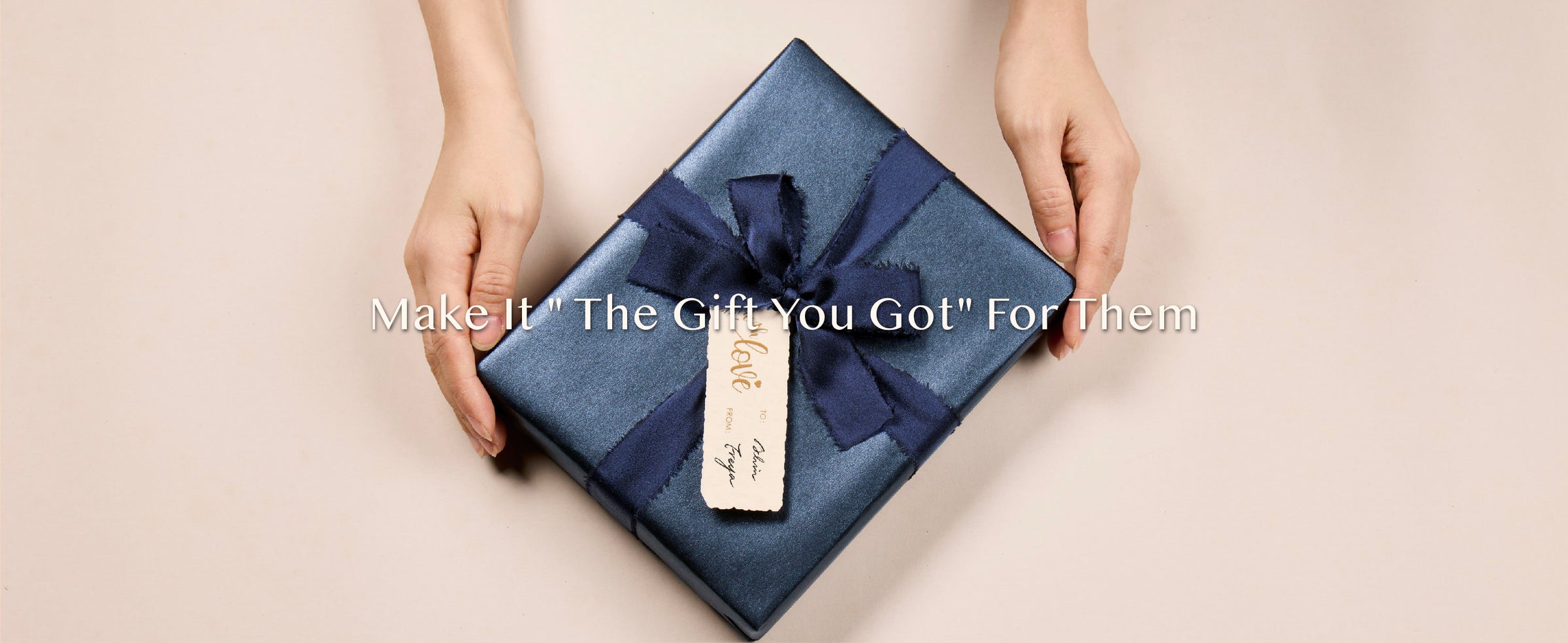 Hands presenting a square gift in shimmering blue paper with a navy bow and cream tag