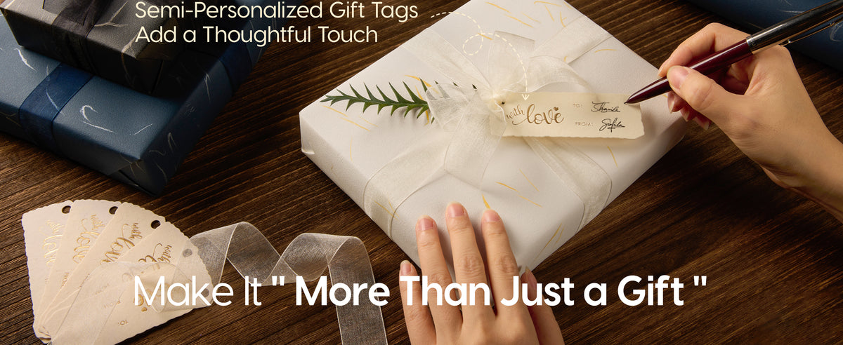 Hands write a gift tag on a white gold-accented package tied with sheer ribbon and greenery.