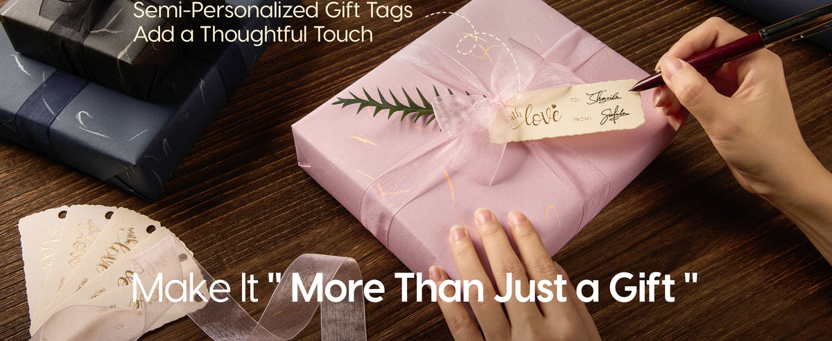 Hands write a gift tag on a pink gold-accented package tied with sheer ribbon and greenery.