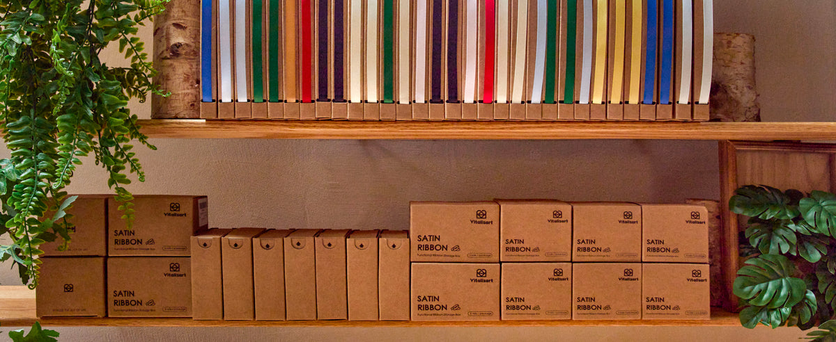 Multicolored satin ribbons in kraft dispensers arranged with storage boxes on warm wooden shelves.