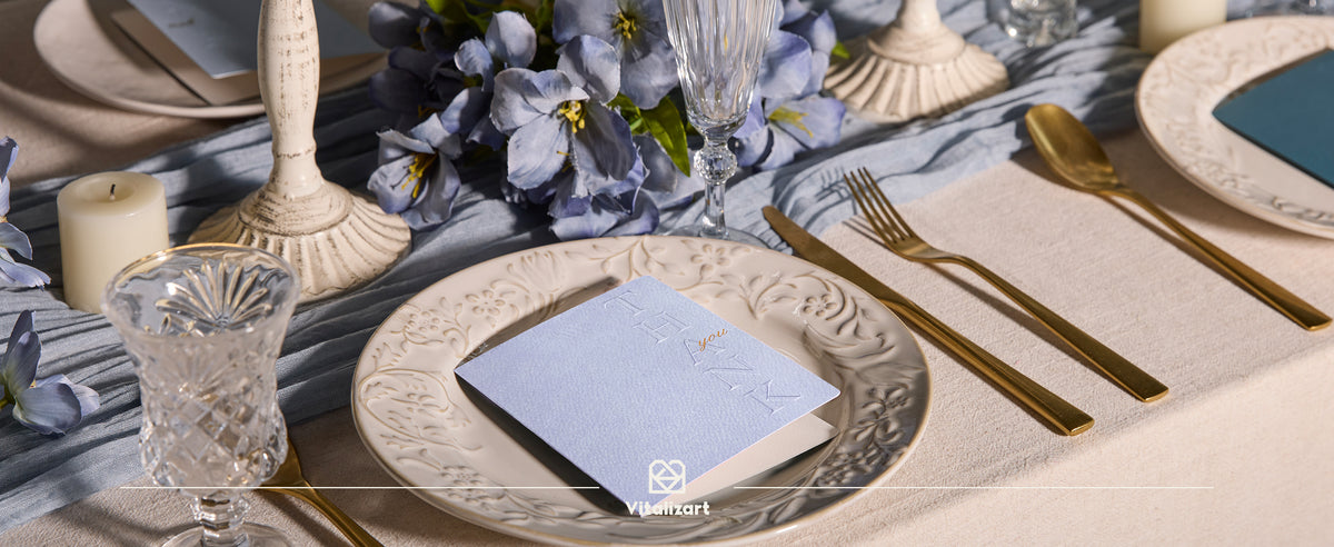 Pale blue-gray embossed thank-you card and kraft envelope on an elegant blue-and-gold table setting