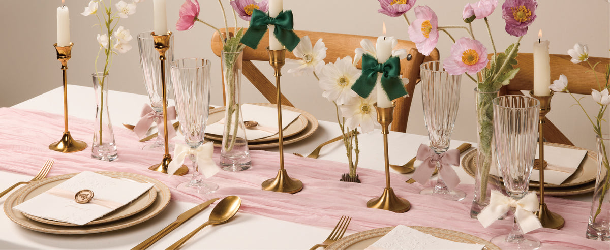 Nude-pink, ivory, and green frayed ribbon bows decorate candles and glasses on a floral dining table.