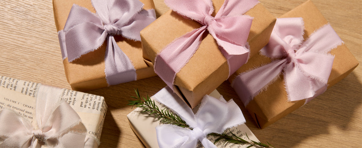 Kraft gift boxes tied with pink and nude frayed satin bows beside a white botanical-wrapped gift.