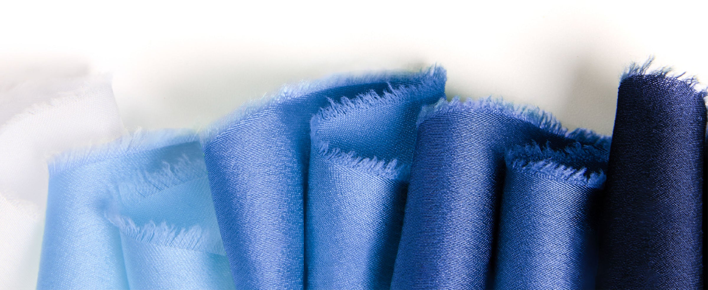Folded frayed-edge satin ribbon samples arranged from navy and blue to pale blue and white.