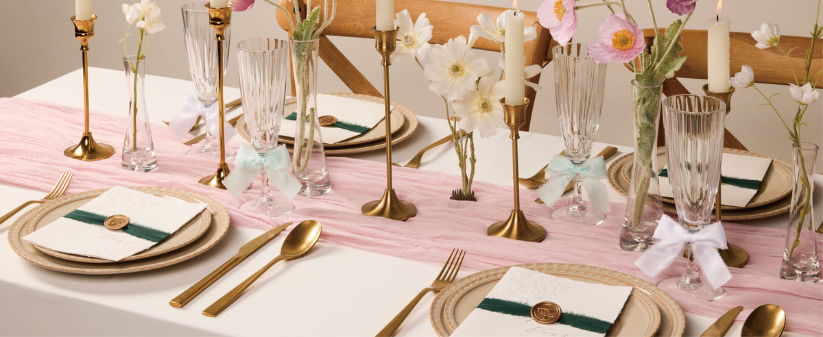 Frayed pastel satin ribbons accent glasses and invitations on a floral table with candles and gold flatware.