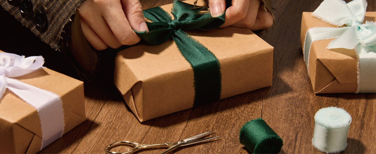 Hands tie dark green frayed satin ribbon around a kraft gift box beside gold scissors and ribbon rolls.