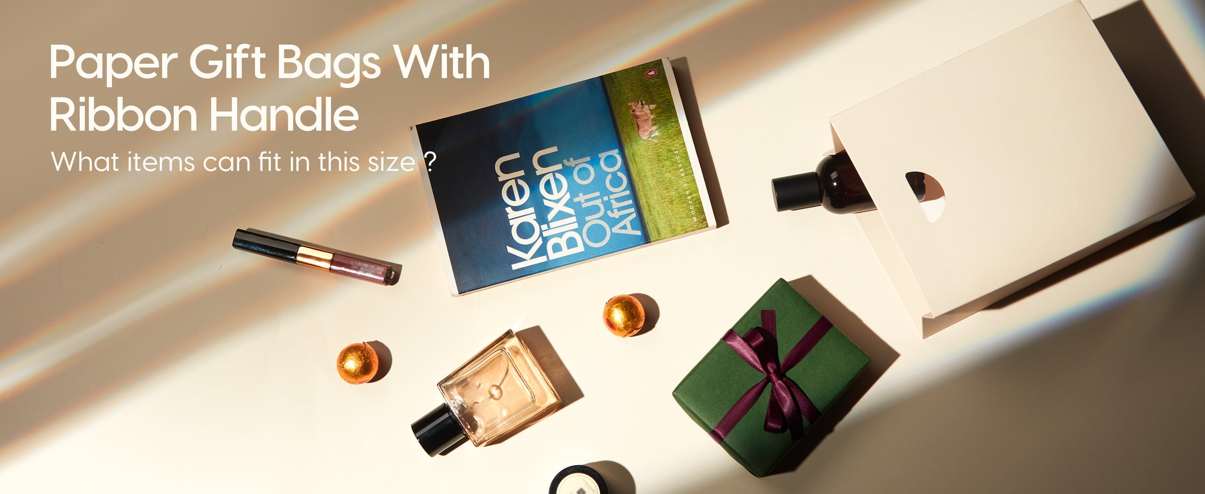 Cream-white paper gift bag beside a book, perfume, candle, cosmetics and a small wrapped gift