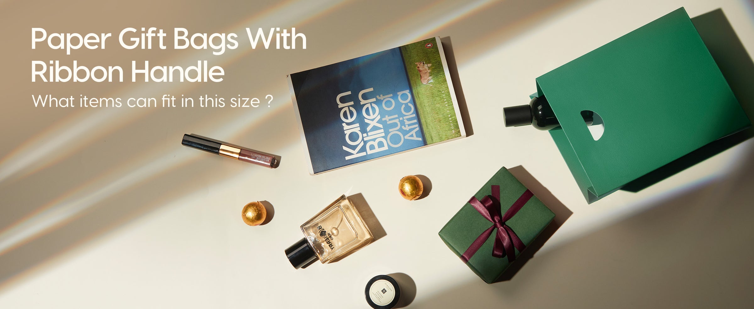 Dark green paper gift bag beside a book, perfume, candle, cosmetics and a small wrapped gift