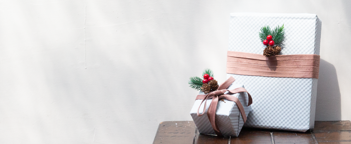 Rose velvet ribbon wraps two white gift boxes accented with pine sprigs, red berries, and pinecones.