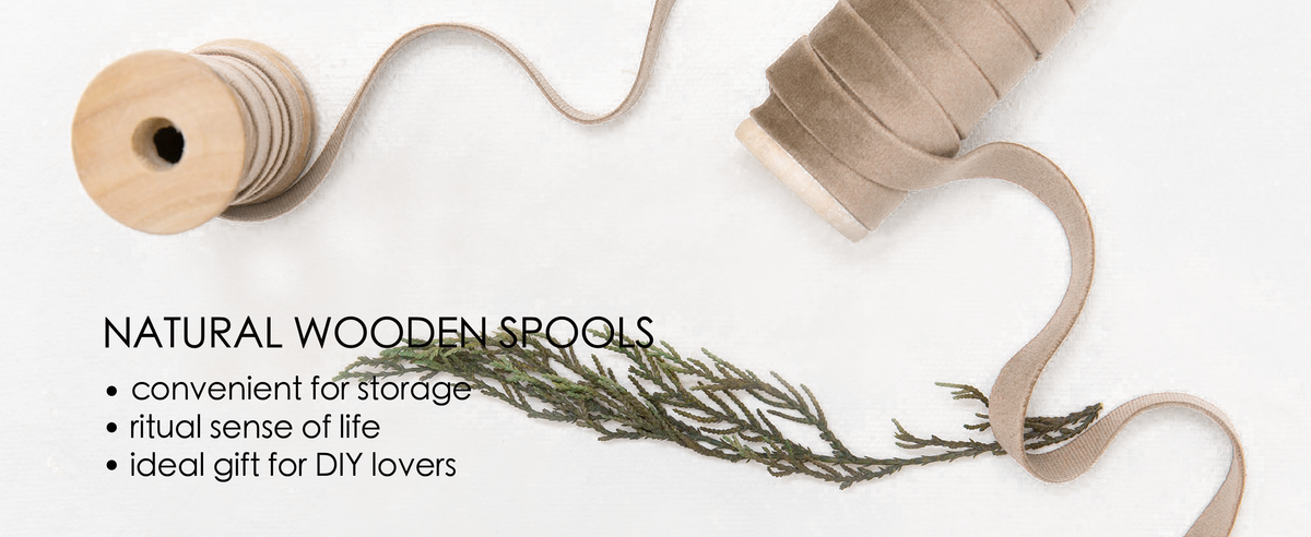 Camel velvet ribbon unwinds between two natural wooden spools on white fabric with greenery.
