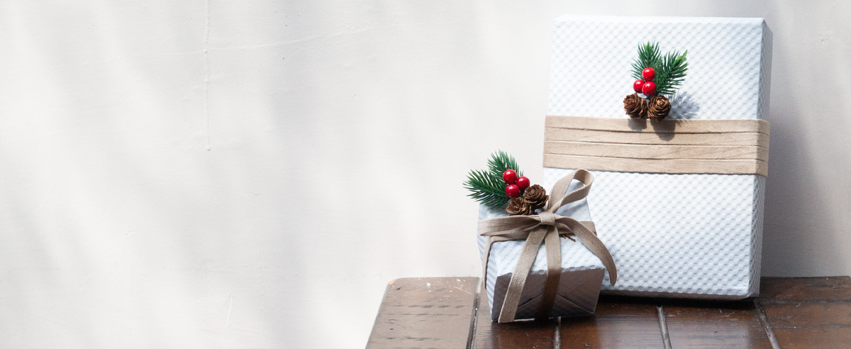 Camel velvet ribbon wraps two white gift boxes accented with pine sprigs, red berries, and pinecones.