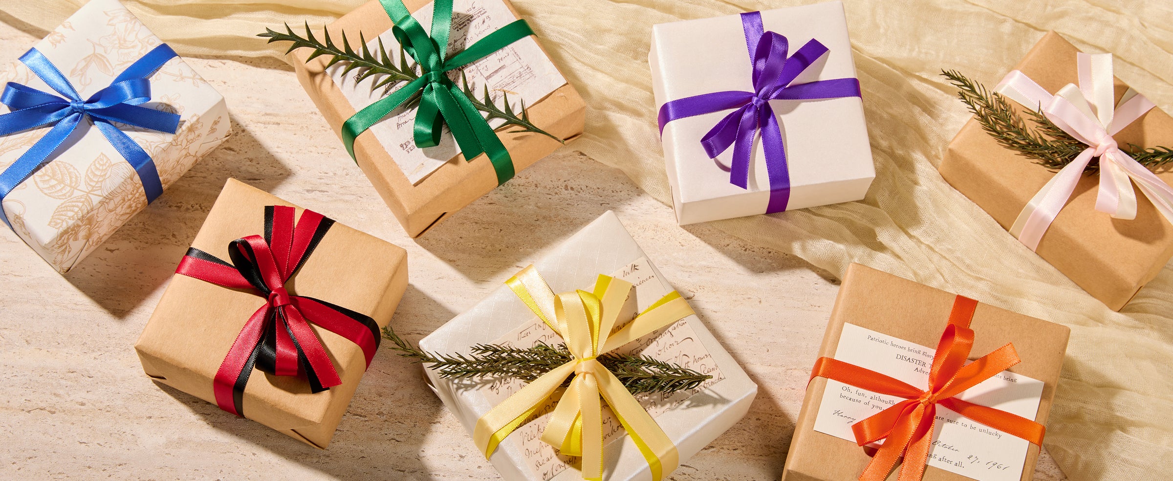 Gift boxes wrapped in kraft and white paper with colorful satin ribbon bows and sprigs of greenery.