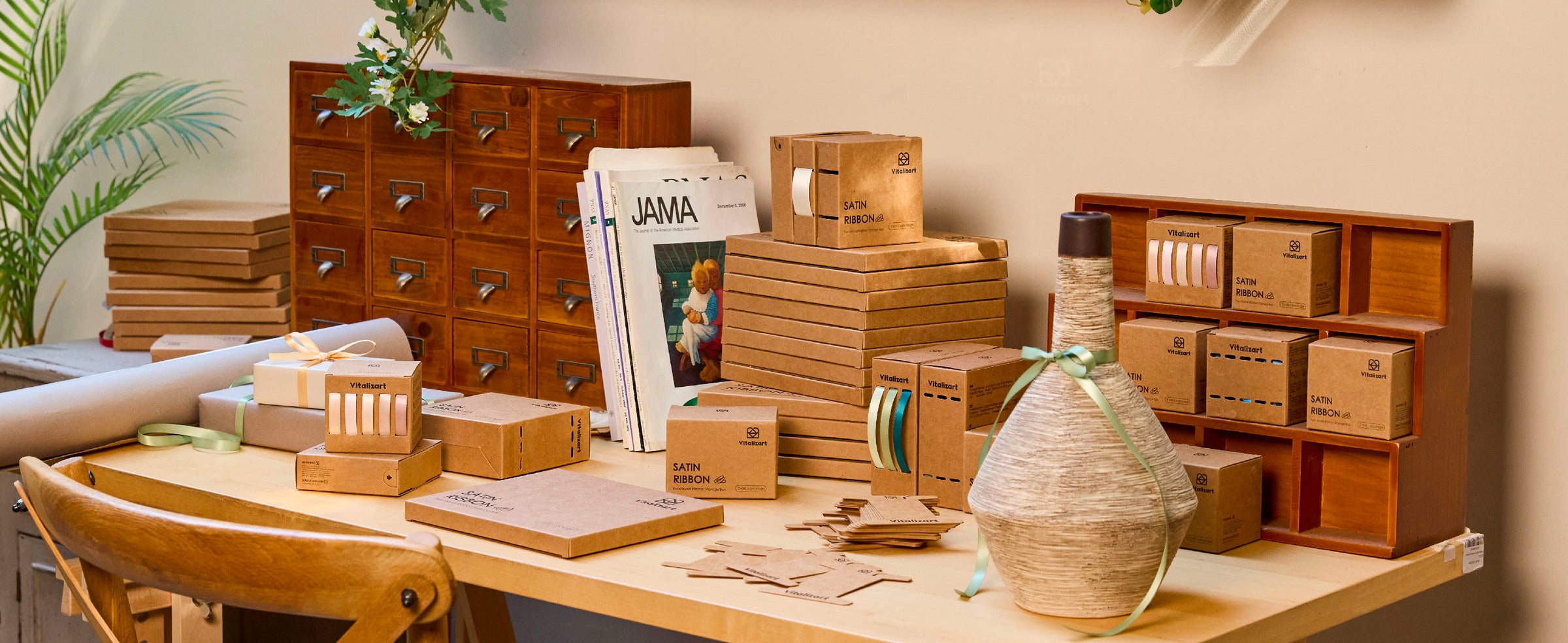 Kraft satin ribbon boxes fill a warm craft workspace with plants, wooden drawers, books, and a bow-tied vase.