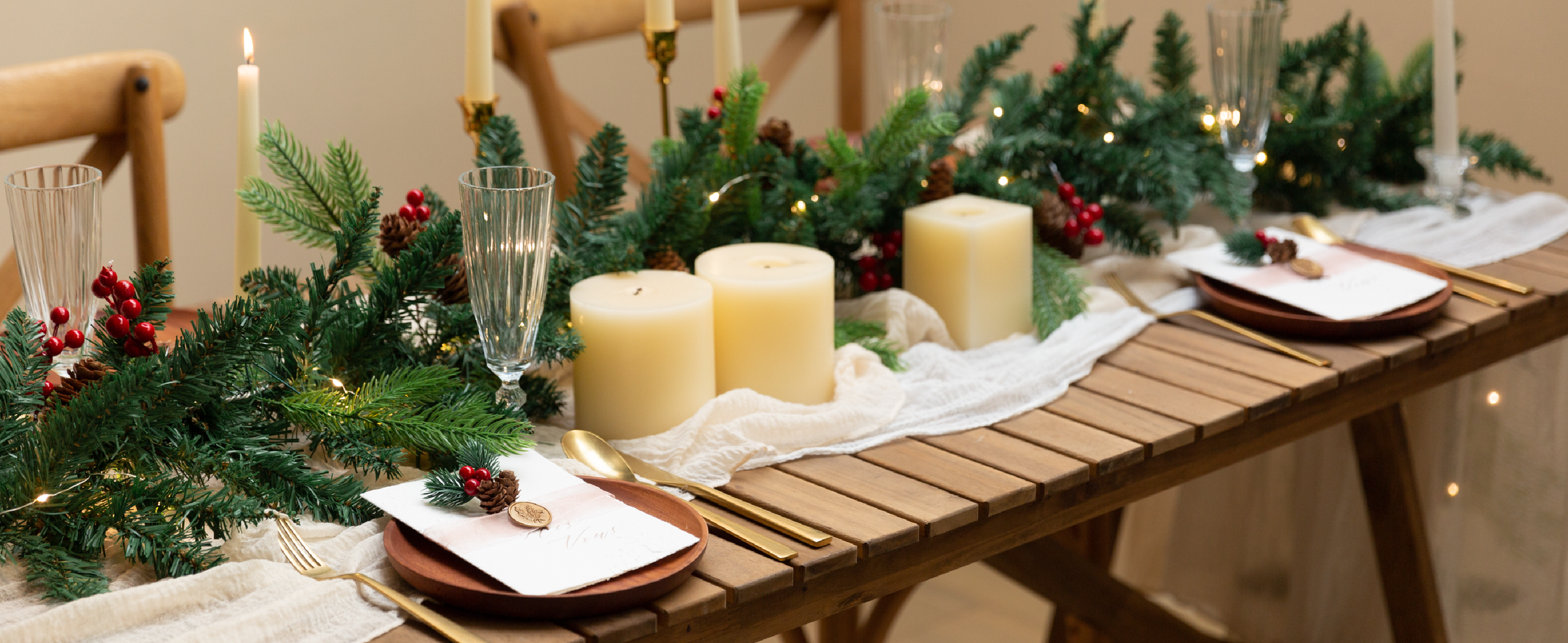 Holiday table with pale ribbon accents, evergreen garland, candles, wooden plates, and gold flatware