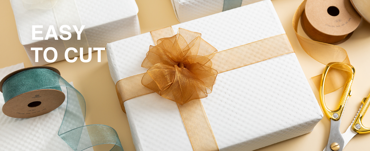 Gold sheer organza ribbon tied into a layered bow on a white gift box beside ribbon rolls and scissors