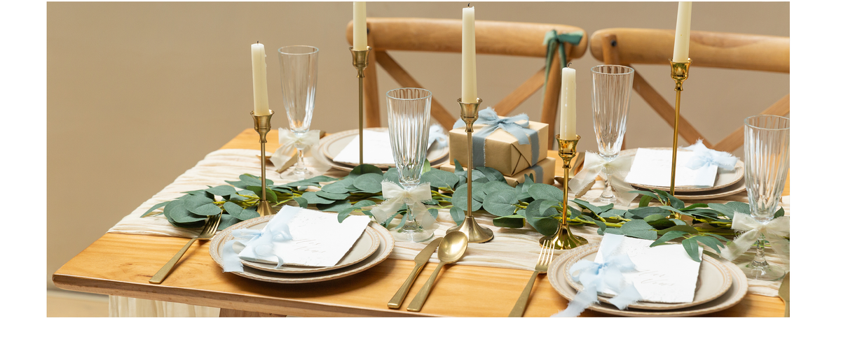 Pale blue and ivory chiffon ribbons accent gifts, glasses, cards, and chairs on a greenery table